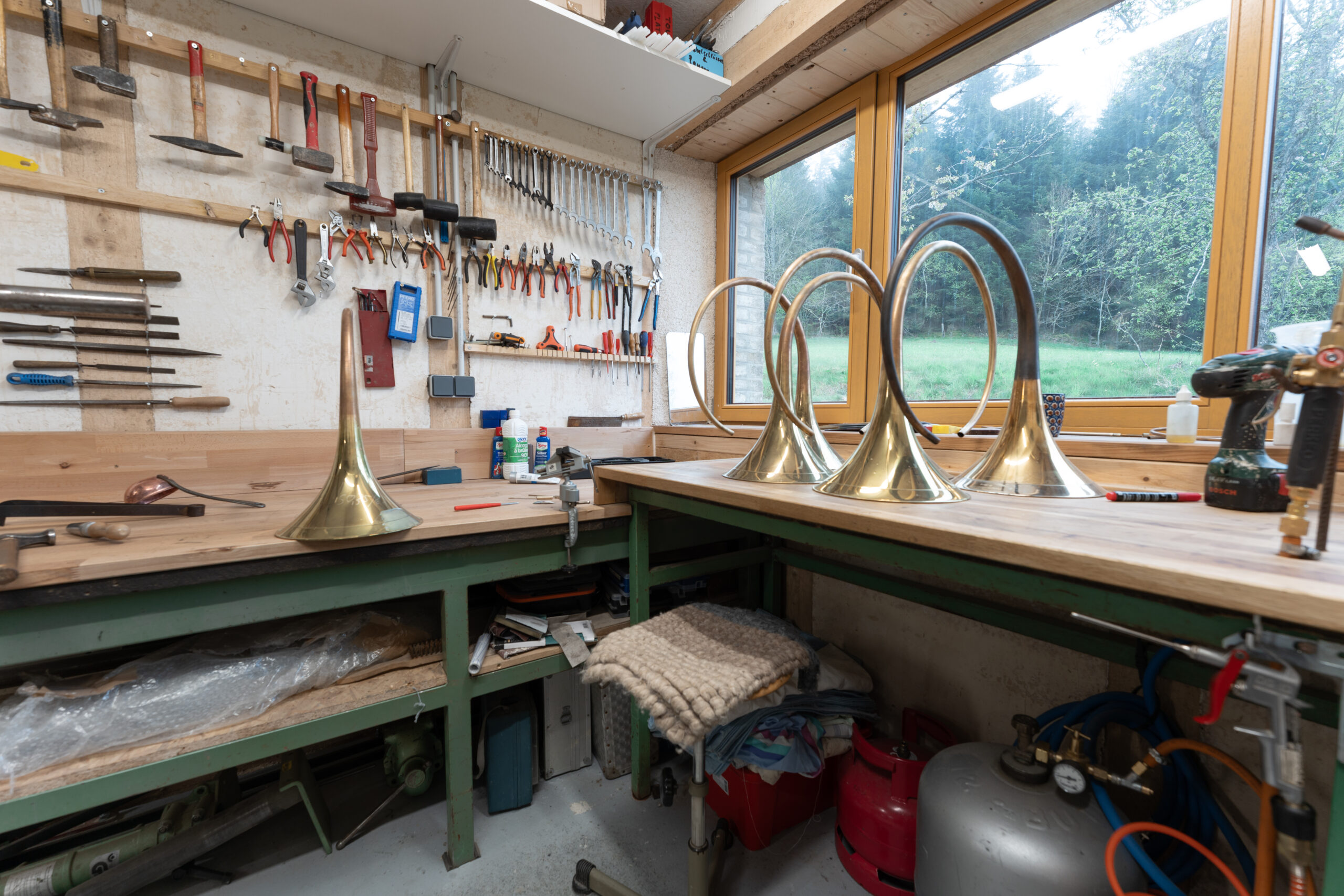Un établi d'atelier avec divers outils accrochés au mur, un pavillon de cor de chasse posé sur le plan de travail, et plusieurs corps de cor de chasse en laiton brillant près d'une grande fenêtre donnant sur la verdure.