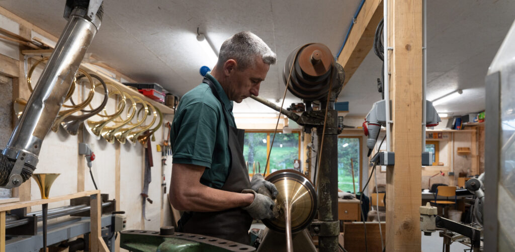Un homme, portant un tablier et des gants, travaille un morceau de laiton brillant sur un tour à repousser dans un atelier. À l'arrière-plan, des pavillons de cor de chasse sont alignés sur une étagère.