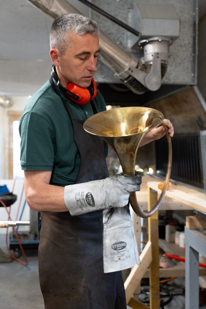 Gérant de Périnet, portant un tablier, des gants de protection épais et un casque anti-bruit autour du cou, examine attentivement une pièce en laiton d'un instrument de musique, probablement un pavillon de cor, dans un atelier.
